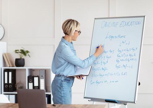 Female university tutor solving complicated math sums during online lesson, explaining logarithms on laptop from home. College professor teaching calculus, giving remote lecture photo