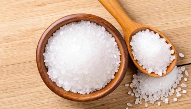 There is a bowl with coarse white salt and a wooden spoon on a wooden table. Several salt crystals are scattered around, creating a harmonious composition. photo
