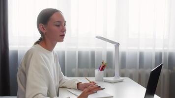 fokuserade studerande flicka skrivning på anteckningsbok medan inlärning uppkopplad använder sig av bärbar dator dator, Sammanträde på vit skrivbord med lampa och färgad pennor nära fönster med gardiner video