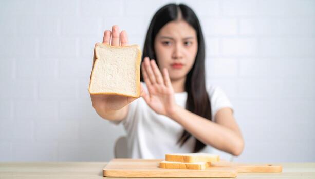 Young woman is rejecting slice of bread, showing clear gesture of disapproval. She appears to be kitchen setting, with wooden cutting board and additional slices of bread nearby. expression her photo