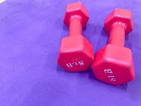 Two red dumbbells are placed on a textured purple mat. The dumbbells are hexagonal with visible weight markings. The lighting is bright and even, highlighting the colors. photo