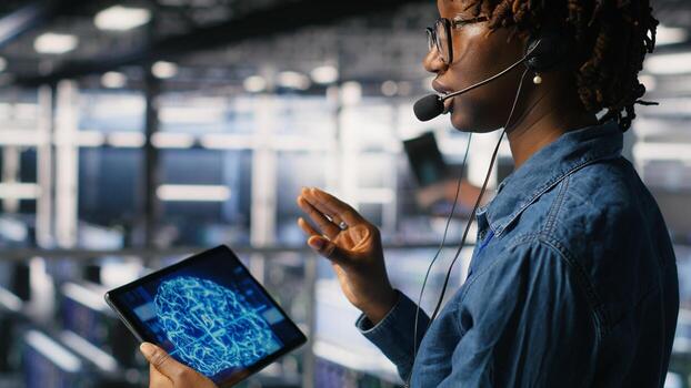 Data center worker on industrial platform speaks in headset microphone while overseeing maintenance on AI rigs below. Admin uses tablet to coordinate deep learning system tasks with team photo