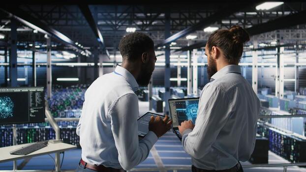 Data center team leader and worker reviewing performance dashboards of AI equipment rigs. Server room supervisor oversees programmer examining machine learning algorithms on screen, photo