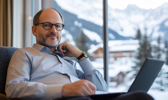 A middleaged man sits in a chair with his hand resting on his chin, looking directly at the camera while using a laptop computer with a snowy mountain view in the background. photo