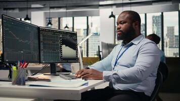 Smiling portfolio manager at computer desk evaluates assets based on statistics and economic indicators. Jolly african american man looks over research reports to drive business growth, camera A video