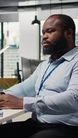 verticale sorridente portafoglio manager a computer scrivania valuta risorse basato su statistica e economico indicatori. gioviale africano americano uomo sembra al di sopra di ricerca rapporti per guidare attività commerciale crescita, telecamera un' video