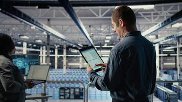 Server room IT expert swiping on tablet touchscreen to read data charts, monitoring equipment health. Admin reviews analysis metrics graphs on device to make sure data center meets goals, camera A video