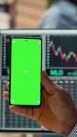 Vertical Trader in prop firm office attends virtual meeting using green screen mobile phone. African american man at work using chroma key mobile phone to participate in internet teleconference, camera B video