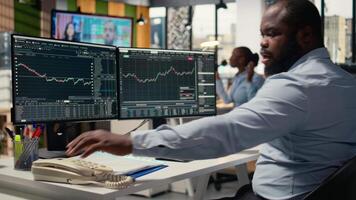 Distressed brokerage firm trader making phone calls to clients in response to Black Swan event shock. African american man evaluate stock exchange impact of unexpected recession, camera A video