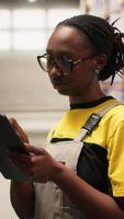 Vertical Black female industrial clerk working on order processing tasks with tablet, distribution center ensures efficient warehousing operation. Managing parcels data for e-commerce delivery. Camera B. video
