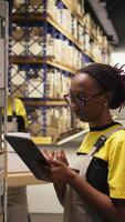 Vertical Black female industrial clerk working on order processing tasks with tablet, distribution center ensures efficient warehousing operation. Managing parcels data for e-commerce delivery. Camera A. video