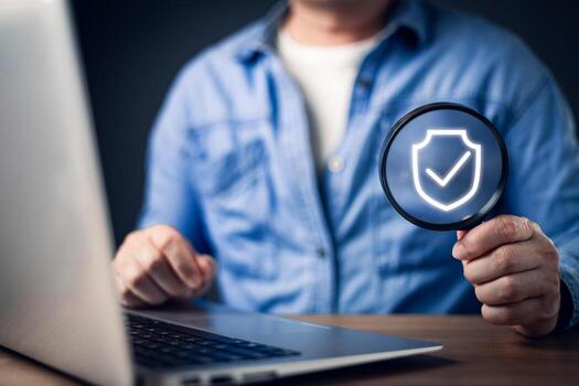 A person in a blue shirt uses a laptop while holding a magnifying glass displaying a glowing shield with a checkmark, symbolizing digital security, data verification, and online protection measures. photo