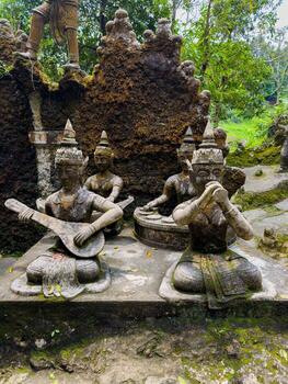 A close-up, medium-shot view of ancient stone statues of mythical musicians, one playing a stringed instrument and another holding a flute-like object, situated in a lush, moss-covered garden photo
