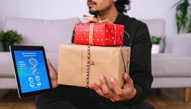 Man sitting floor holding two wrapped gift boxes, one with red wrapping paper and bow, other with plain brown paper and string, cozy living room setting with tablet showing DNA helix graphic photo