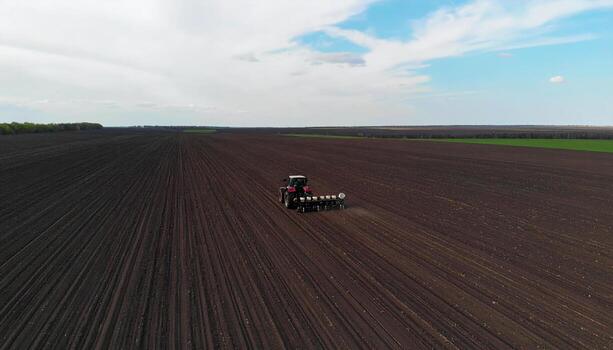 Aerial view of tractor working on large farmland with dark soil and green patches in distance under partly cloudy sky, capturing essence of agriculture and farming activity photo