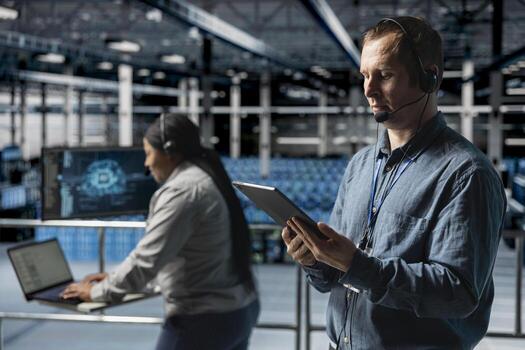 Data center admin wearing headset microphone providing remote diagnostics using tablet. Technician monitors system health and guides users through infrastructure troubleshooting steps photo