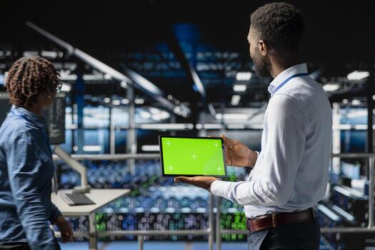 Data center programmer on industrial platform using green screen tablet, solving optimization tasks around the server farm. Male worker visualizing system performance using mockup screen. photo