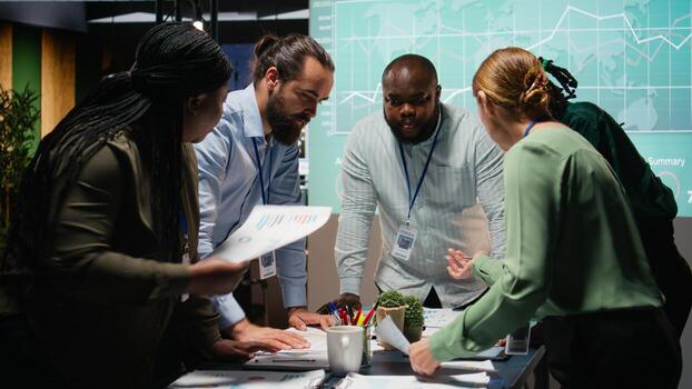 Multiethnic team exchanging files and review forecasting diagrams at night, meeting in a quiet office space. After hours collaboration to prepare key project updates for shareholders. Camera A. photo