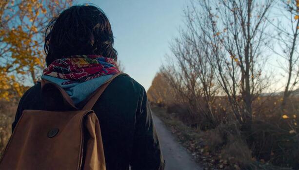 A person is walking down a path with a backpack on. The backpack is brown and has a red scarf tied around it. The person is wearing a black jacket and scarf. The scene is peaceful and serene photo