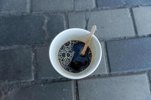 un desechable taza lleno con humeante café se sienta en un guijarro camino, atrapando luz de sol. un de madera agitador descansa adentro, reticente a un reciente verter foto