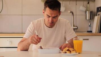 uma homem senta às uma luz cozinha mesa, comendo uma forte refeição enquanto bebericando refrescante laranja suco. a atmosfera é brilhante e convidativo, exibindo uma moderno culinária espaço. video