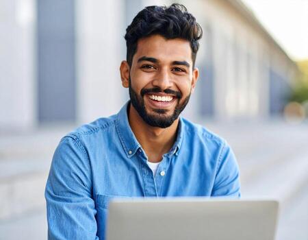 Young man smiling while using laptop outdoors, showcasing joyful expression and casual attire. setting reflects modern environment, perfect for work or leisure photo