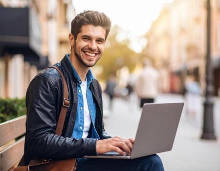 Young man smiling while using laptop outdoors, showcasing relaxed and happy demeanor. scene captures vibrant urban atmosphere with people in background photo