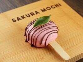Delicious pink mochi treat drizzled with chocolate on a wooden plate, garnished with a leaf. photo
