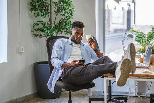 A relaxed man sitting cross-legged on the table using his smartphone in a modern coworking space in front of the computer. photo