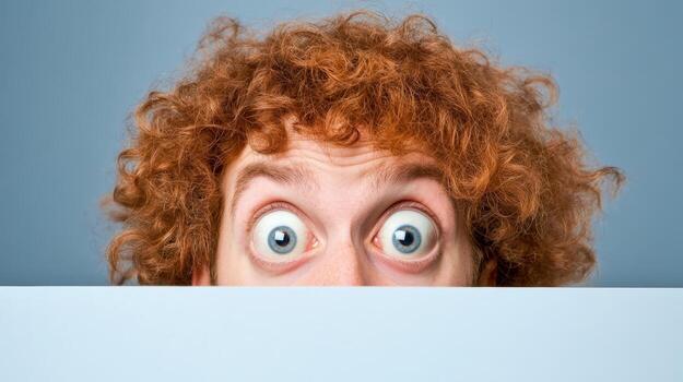 Surprised man with curly hair peeking over a blank surface in a neutral setting photo