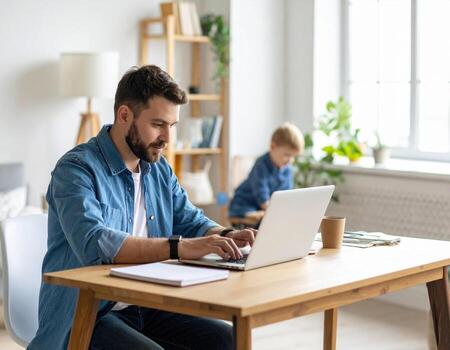 Man working on laptop at wooden table with notebook and coffee cup, child playing in background near window, bright home office with natural light and plants photo