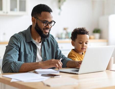 Man working on laptop with child sitting beside him at home, showing warm and focused atmosphere of remote work and family bonding in bright kitchen setting photo