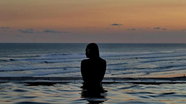 Back view. Silhouette of Woman in Infinity Pool Watching Ocean Sunset with Crowded Beach and Colorful Sky photo