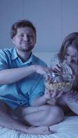 afetuoso casal carinho, assistindo filme, partilha Pipoca tigela, rindo suavemente dentro acolhedor quarto durante romântico noite em uma rainha Tamanho cama capturado dentro vertical modo video
