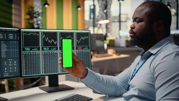 Trader in prop firm office attends virtual meeting using green screen mobile phone. African american man at work using chroma key mobile phone to participate in internet teleconference, camera A video