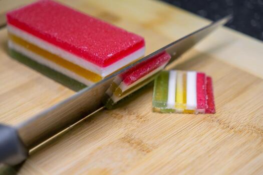 Vibrant multicolored layered jelly candy, coated in sugar, being sliced with a sharp knife on a rustic wooden board, creating a sweet dessert photo