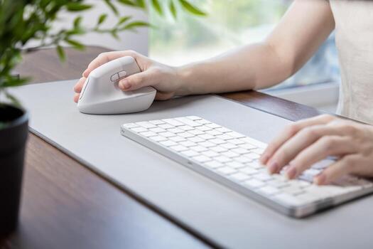 Person's hand operates a modern ergonomic vertical mouse and keyboard on a desk pad, emphasizing comfort and efficiency for prolonged remote work photo