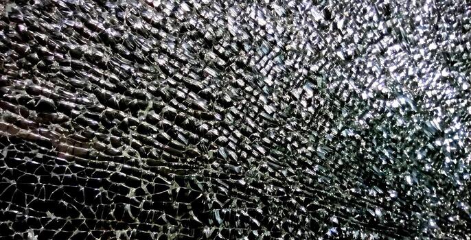 The photo captures a close-up of shattered glass, showcasing an intricate network of sharp fragments reflecting light. The dramatic texture and gleaming surfaces create a striking visual effect.