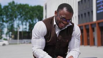 zoom ut kamera se av de eleganta attraktiv man Sammanträde på de bänk och använder sig av hans smartphone. har skägg och eleganta glasögon. han leende med glädje video