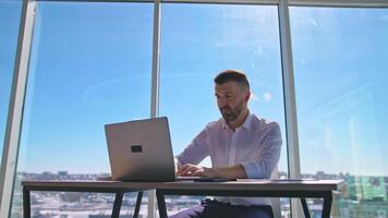 focado homem de negocios trabalhando em computador portátil computador dentro escritório com ampla janelas. barbudo masculino trabalhador usando computador portátil computador às ambiente de trabalho. bonito homem olhando às computador portátil tela dentro escritório video