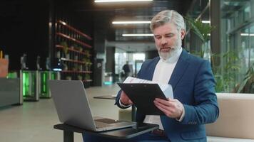 Lycklig affärsman använder sig av bärbar dator och arbetssätt med dokument, analyserar information, läsning papper förbereda finansiell Rapportera. manlig verkställande i kostym stängning netbook efter efterbehandling arbete i kontor lobby. video