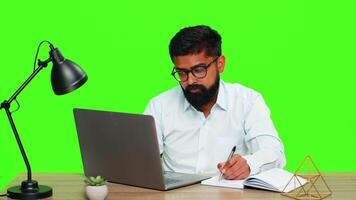 indisk man sitter på skrivbord skrivning idéer i anteckningsbok medan arbetssätt på bärbar dator, som visar fokus, planera, göra flera saker samtidigt. arab kille isolerat på grön krom nyckel bakgrund uttrycker produktivitet tänkande. video