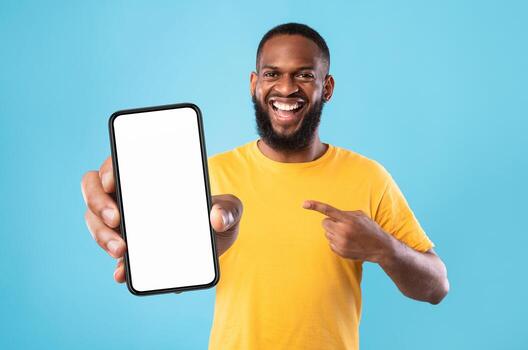 Handsome happy black guy pointing at smartphone with blank screen on blue studio background, mockup for website or new mobile app. Cellphone display template. Space for advertisement photo
