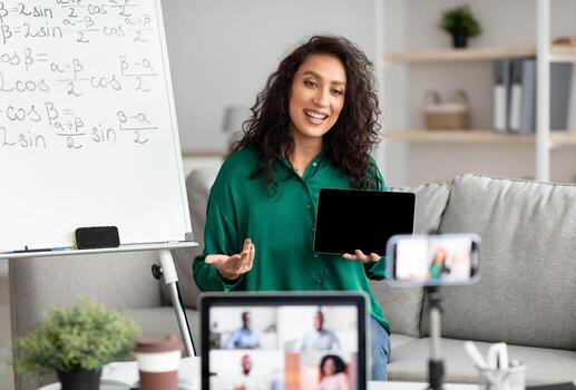 Female math teacher making conference chat with diverse group of students using laptop and cellphone on tripod, holding showing tablet with empty screen with copy space for mockup, explaining photo