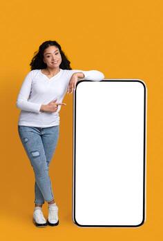 Smiling African American Lady Leaning And Pointing At Big Smartphone With Blank White Screen, Showing Copy Space For Your App Or Website Design, Standing Over Yellow Studio Background, Mockup Image photo