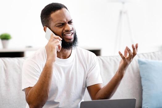 Discontented Black Guy Talking On Cellphone Sitting At Laptop Having Computer Problem At Home. Displeased Customer Calling To Helpline Having Issue With Internet Connection Concept. Selective Focus photo