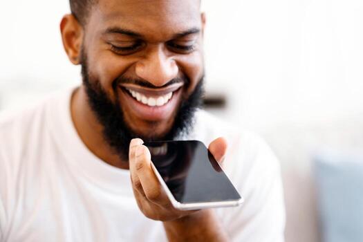 Closeup Of African Guy With Phone Using Voice Assistant App Talking Holding Smartphone Near Mouth Indoors. Man Chatting Using Loudspeaker, Browsing Internet Via Voice Search App. IOT Smart Technology photo