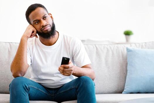 Bored African Man Watching TV Switching Television Channels With Remote Control Sitting On Couch At Home. Discontented Viewer, TV Programming Problem Concept. Free Space For Text photo