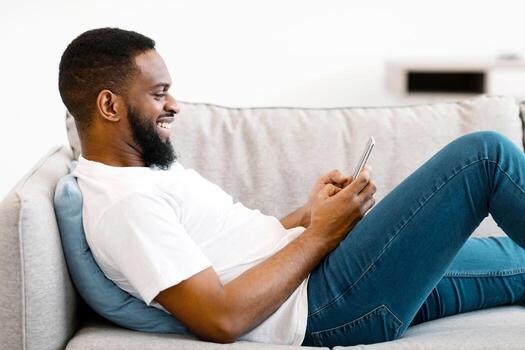 Great Mobile App. African Man Using Cellphone Texting And Browsing Internet Lying On Couch At Home. Guy Networking In Social Media Application Using Smartphone. Side View photo