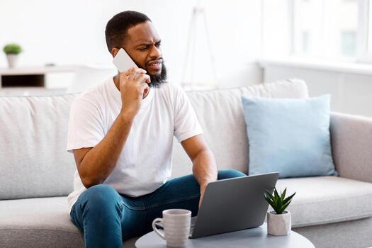 Displeased African Man Talking On Mobile Phone Sitting At Laptop Having Problem With Customer Support Helpline At Home. Discontented Client And Computer Issue Concept. Selective Focus photo